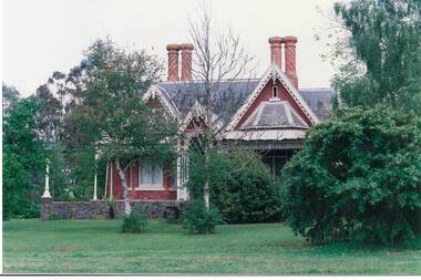 Front view tall brick house in high stone foundation, high orate gables, partially blocked by trees.