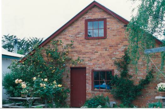 End of brick building, peaked roof with loft window, window and door (ïndian" red), lemon tree and roses on left, birch tree on right.