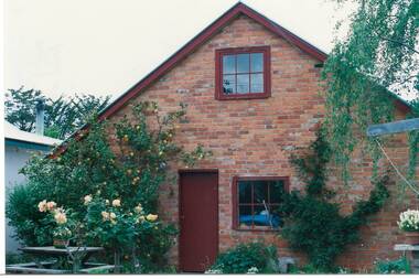End of brick building, peaked roof with loft window, window and door (ïndian" red), lemon tree and roses on left, birch tree on right.