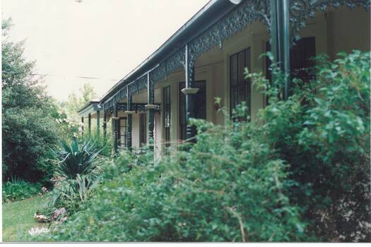 Looking through lush garden along front veranda, wooden posts with ornate iron lace decorations, painted Brunswick Green.