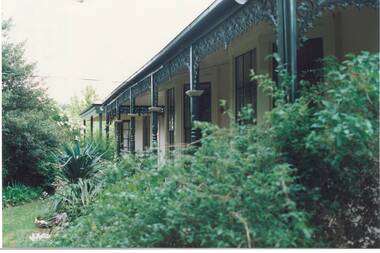Looking through lush garden along front veranda, wooden posts with ornate iron lace decorations, painted Brunswick Green.