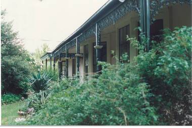 Looking through lush garden along front veranda, wooden posts with ornate iron lace decorations, painted Brunswick Green.