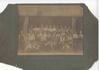 Three rows of workers standing, with another four workers seated on the ground, in front of large dark shed. 