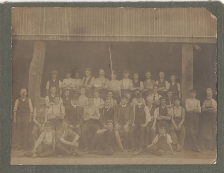 Three rows of workers standing, with another four workers seated on the ground, in front of large dark shed. 