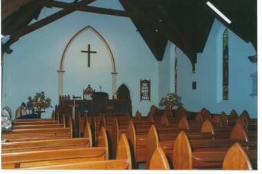 Interior of church, white walls, wooden pews, Flowers either side of communion table, Hymn board.
