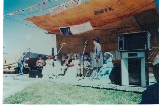 Group of people in period Victorian dress under canvas shelter, large speakers on right.