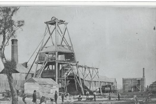 Mine structures including wooden poppet, sheds, two brick chimneys, railway engine and freight wagon, men and a dog middle distnce.