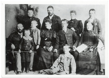 Family group, four grown children standing at rear, parents seated either side of three younger children, another child seated on floor, and youngest on fathers lap, all in Victorian dress.
