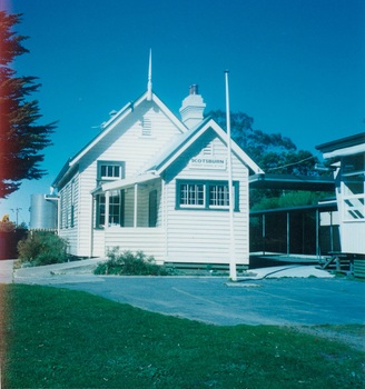 White woden school building, high peaked roof, small extended section at front with verandah