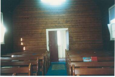 Interior of church, unpainted wood-lined walls, door centre, rows of pews.