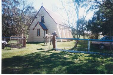White weatherboard church, high pitched roof, chain link fence and gates, trees either side.