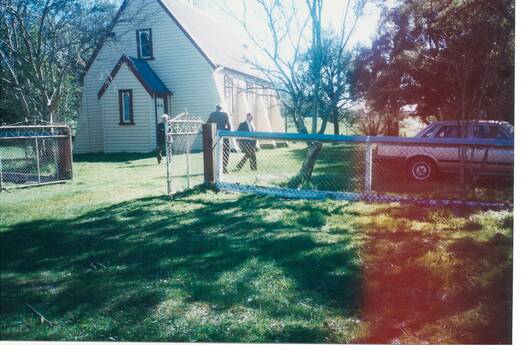 White weatherboard church, high pitched roof, chain link fence and gates, trees either side.