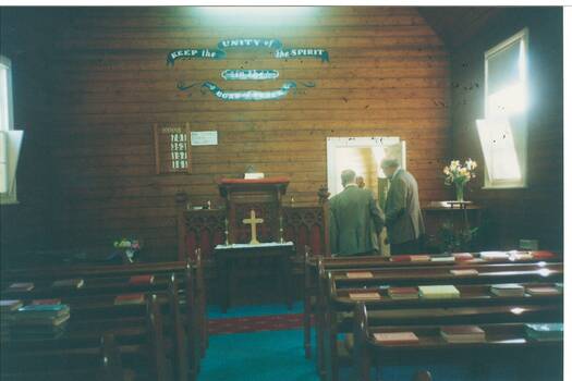 Interior church, unpainted wood-lined walls, communion table, pews with hymnals