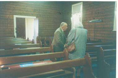 Interior wood-lined church, open door at rear, pews, lady and gentleman standing in aisle.