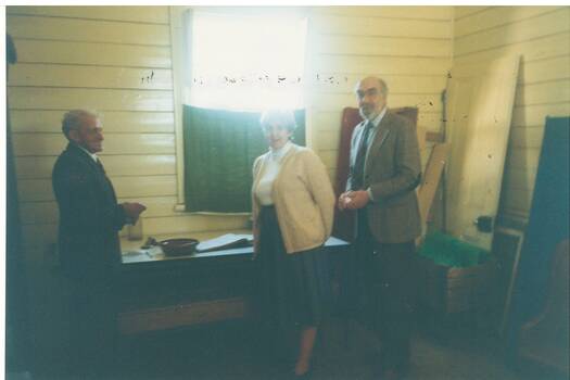 Interior, white painted wood lined walls, widow with half curtain, lady and two gentlemen standing centre.