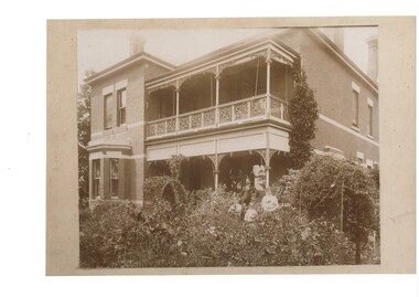 Two story brick house, veranda with iron lace, established garden, Couple with two children in garden.