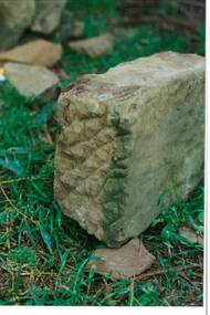 Rectangular sandstone block, rough hewn, chisel marks very obvious, on grass, rock fragments in background.