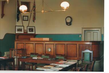 Cream walls with green dado, looking over tables to raised Judge's bench, hanging brass lamps.