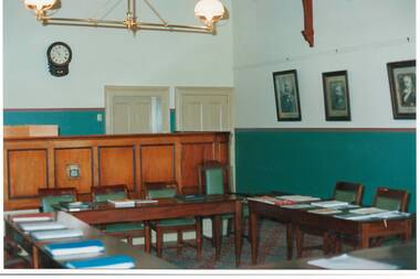 Corner of room, cream walls with green dado, panelled raised Judge's Bench, tables and chairs, portraits on wall.