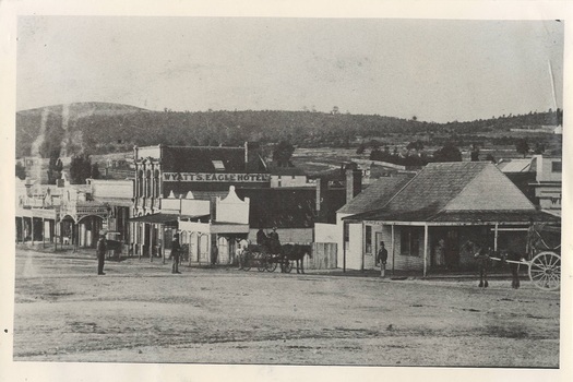 Street scene, single story Victorian shops, two story brick hotel centre, treed hills behind. Some men middle distance, and two carts with horses.