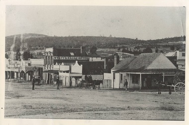 Street scene, single story Victorian shops, two story brick hotel centre, treed hills behind. Some men middle distance, and two carts with horses.