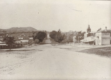 View up tree-lined dirt road towards Mount, shops along left side, more substantial buildings on right.