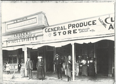 Two stores with verandah, signage for Produce, wine & spirits, grocery. People standing in front, goods visible on footpath and in windows.