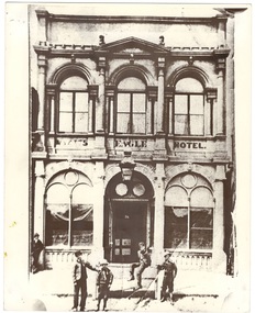 Two story, stucco hotel, arched entrance flanked by arched windows, three arched windows above, all separated by columns, pediment above. boys in front.