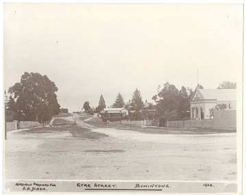 Streetscape, wide dirt road, Public Hall on right, houses far centre, and trees and picket fence left.
