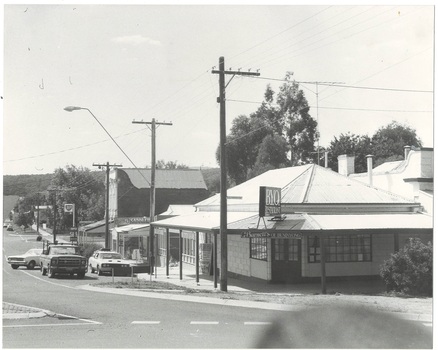 Streetscape across intersection, single story corner shop with iron roof on corner, taller brick building to right, street running out to left shops and service station.