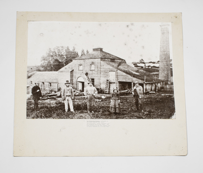 Black and white photograph of five people standing in front of a bluestone, brick and timber building with a brick chimney nearby.