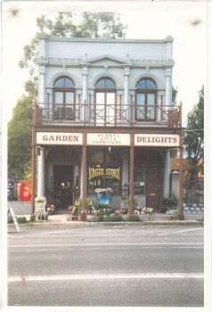 Narrow 2 story building, white stucco, veranda, sign "Garden Delights" first floor arched window either side of arched doorway, pillars between and pediment above.