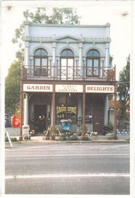 Narrow 2 story building, white stucco, veranda, sign "Garden Delights" first floor arched window either side of arched doorway, pillars between and pediment above.