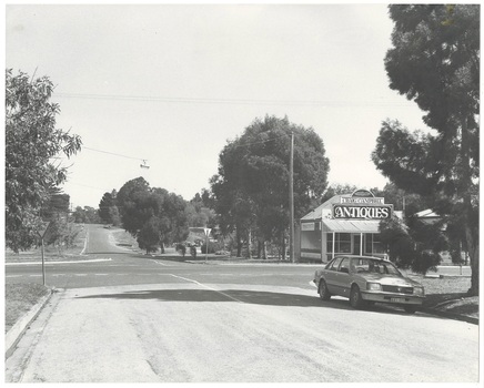 Streetscape along road over intersection, small antique shop on right corner, parked car, trees along street.