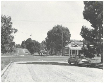 Streetscape along road over intersection, small antique shop on right corner, parked car, trees along street.