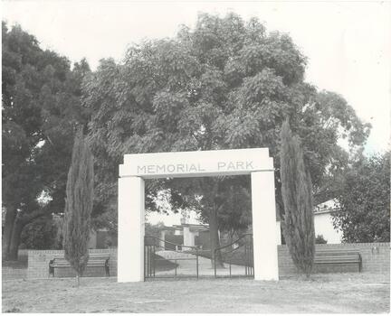 White block archway, inscribed "Memorial Park," black iron gates, brick half walls, cypress trees each side, and large trees in park.