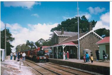 Bluestone railway station, slate roof, steam train centre, people on platforms both side of rails.