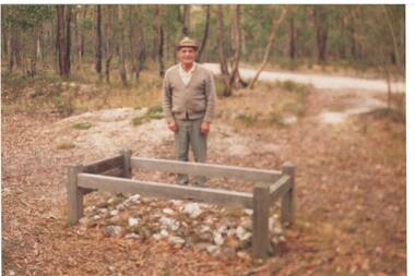 Man standing behind grave with neat wooden surround, in forest clearing, dirt road running behind.