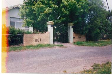Tall rendered posts with iron gates, lower brick wall either side   inscribed 1864 and 1964, weatherboard building behind.