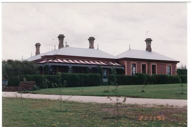 L-fronted polychrome brick homestead, tin roof, four chimneys, wrap-around veranda, painted red stripes, left wing, hedge across front, lawn and path foreground.