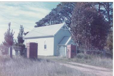 Small wooden church, peaked tin roof, entry annex, gravel drive with more modern iron gated and brick pillars.