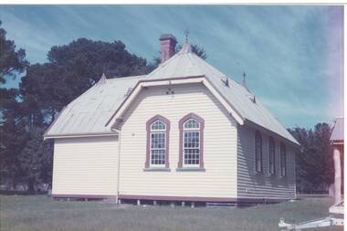 Colour Photograph of South Face of school. Wooden cream painted building, with iron roof. Roof points decorated with air vents. Two school arched windows with multiple small panes, painted in burgundy.