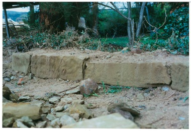 Row of sandstone blocks at ground level,  rubble in foreground, rough garden behind.