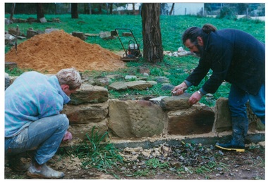 Two men laying sandstone blocks for wall, grass, trees and stone blocks in background.