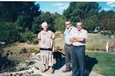 Lady and two men standing in garden, sunny day, neat paths and garden beds, trees in backgrouond