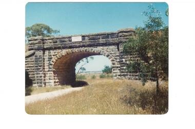 Colour photograph of bluestone railway bridge showing arch and unmade road underneath arch