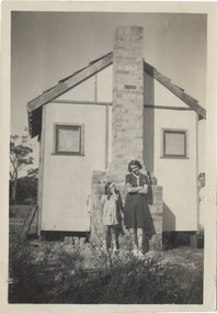 End-on view of shed, with brick chimney, small window either side. Two girls leaning on chimney, another peeking around right side.