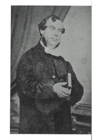 Portrait of middle aged man wearing clerical garments, hands clasped holding bible at chest level, darpes in background.