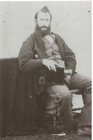 Portrait of bearded man in Victorian clothes, loose jacket, waistcoat, narrow-striped trousers, holding early camera resting on his leg.