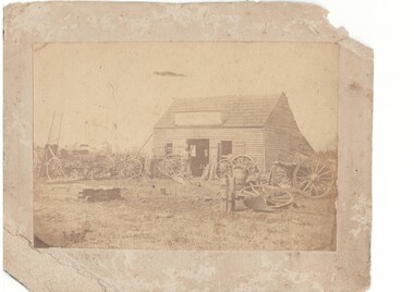 Simple wooden building with shingle roof, central open double door flanked by unglazed windows with shutters, surrounded by wagons, wheels, ploughs etc.